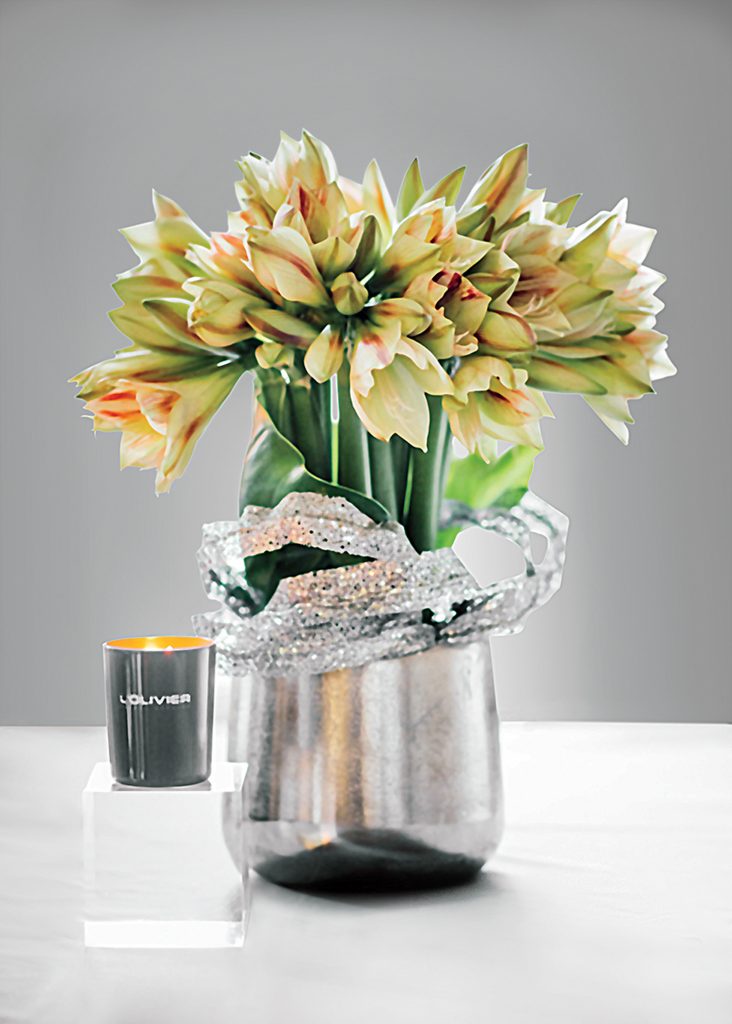 A sparkling twist encircles a cluster of striped amaryllis, One of L’Olivier’s scented candles completes the presentation.